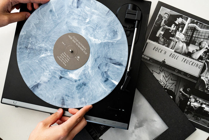 A person places a blue and white marbled Cloud Nothings vinyl record on a turntable, surrounded by black and white album covers, photos, and a visible tracklist featuring “ROCKN ROLL TRUCKING.”.