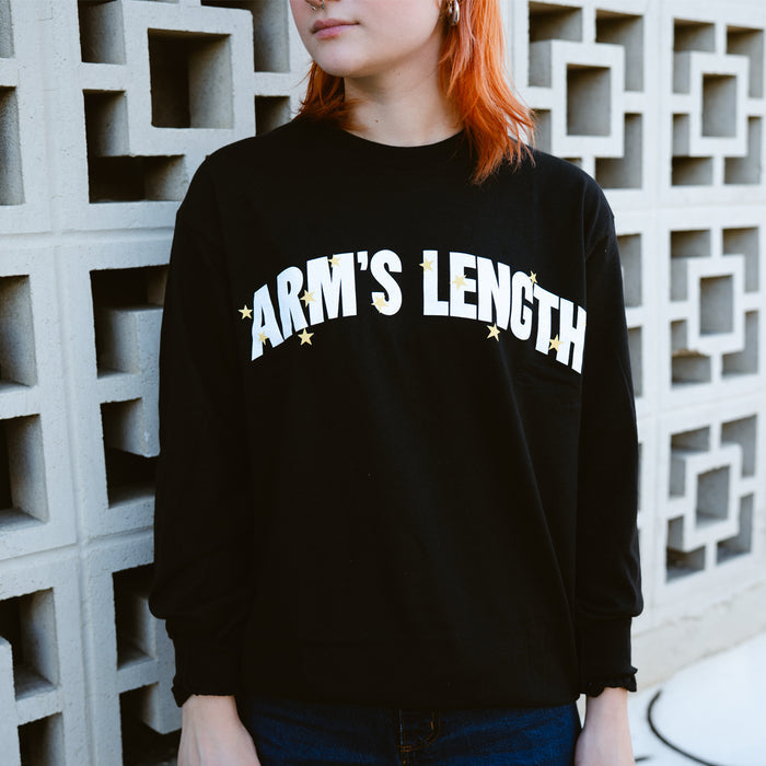 A person with shoulder-length orange hair wears a black Arms Length long-sleeve shirt with ARM’S LENGTH in white letters and small yellow stars, standing before a geometric concrete wall.