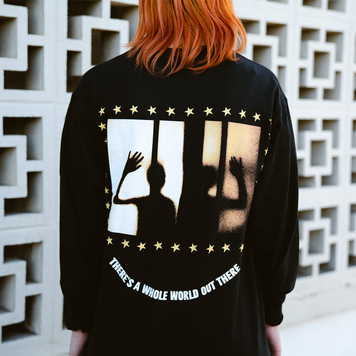 A person with shoulder-length red hair wears an Arms Length black shirt featuring silhouetted figures waving from windows, yellow stars, and the text “THERE’S A WHOLE WORLD OUT THERE.”.