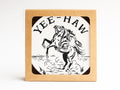 The Yeehaw Cowgirl Coaster Set by Onderkast Studio comes in a square window box featuring a cowgirl on a rearing horse with YEE-HAW above—a fun way to showcase these handmade letterpress coasters.