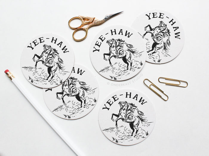 The Yeehaw Cowgirl Coaster Set by Onderkast Studio includes five round, recyclable letterpress coasters featuring a black cowboy on a bucking horse and YEE-HAW text. Gold scissors, paperclips, and a white pencil with red eraser are nearby.